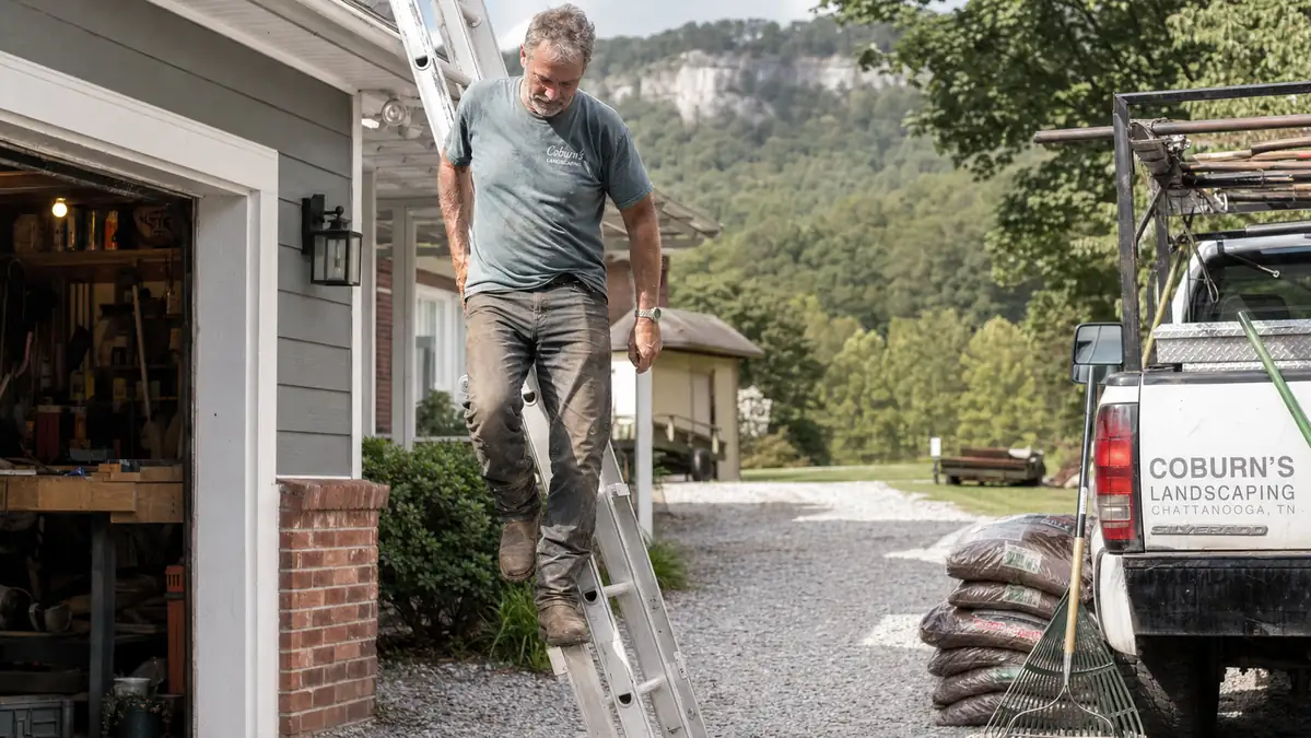Landscaper climbing down the last rungs of a ladder after clearing gutters, work truck and mulch bags visible nearby