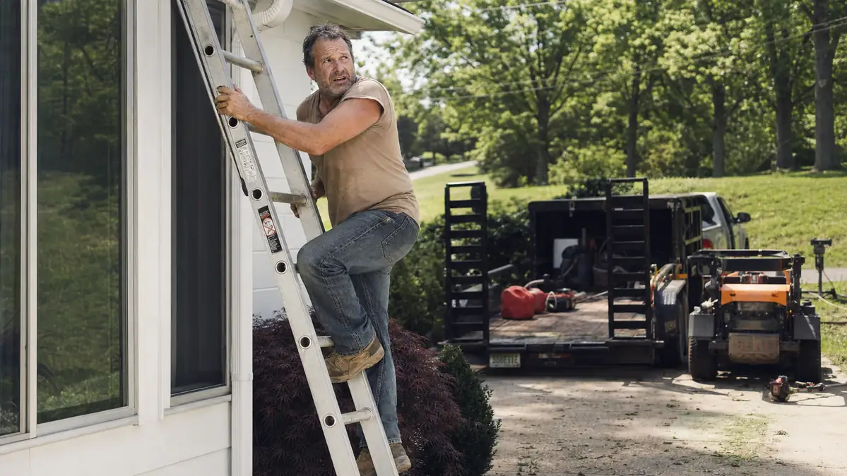 Landscaper climbing an extension ladder against a house with a mower and trailer visible in the driveway